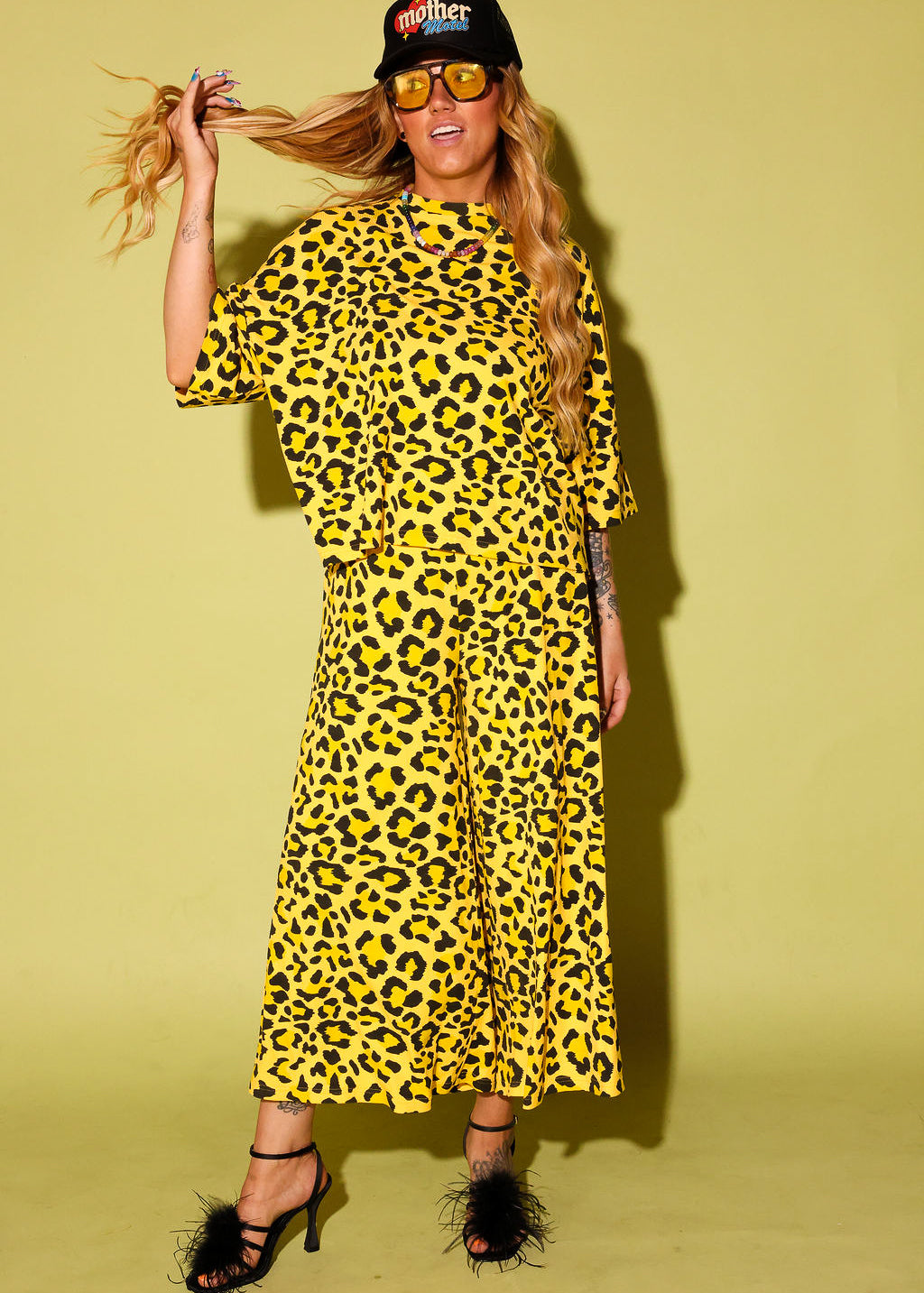 Model wearing a black and yellow leopard set black high heels and a black trucker hat that says 'mother' on it, against a pale yellow background