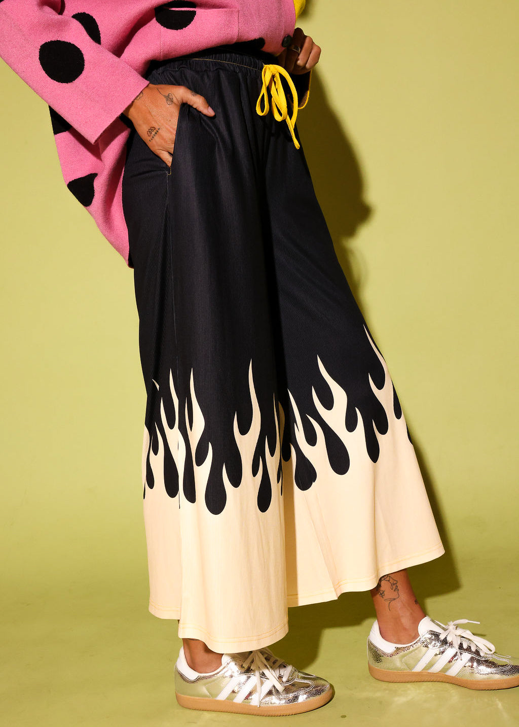 A person wearing wide-leg pleated pants with a black upper section and a cream lower section featuring a black flame print, styled with a pink polka-dot sweater and metallic sneakers against a yellow-green background.