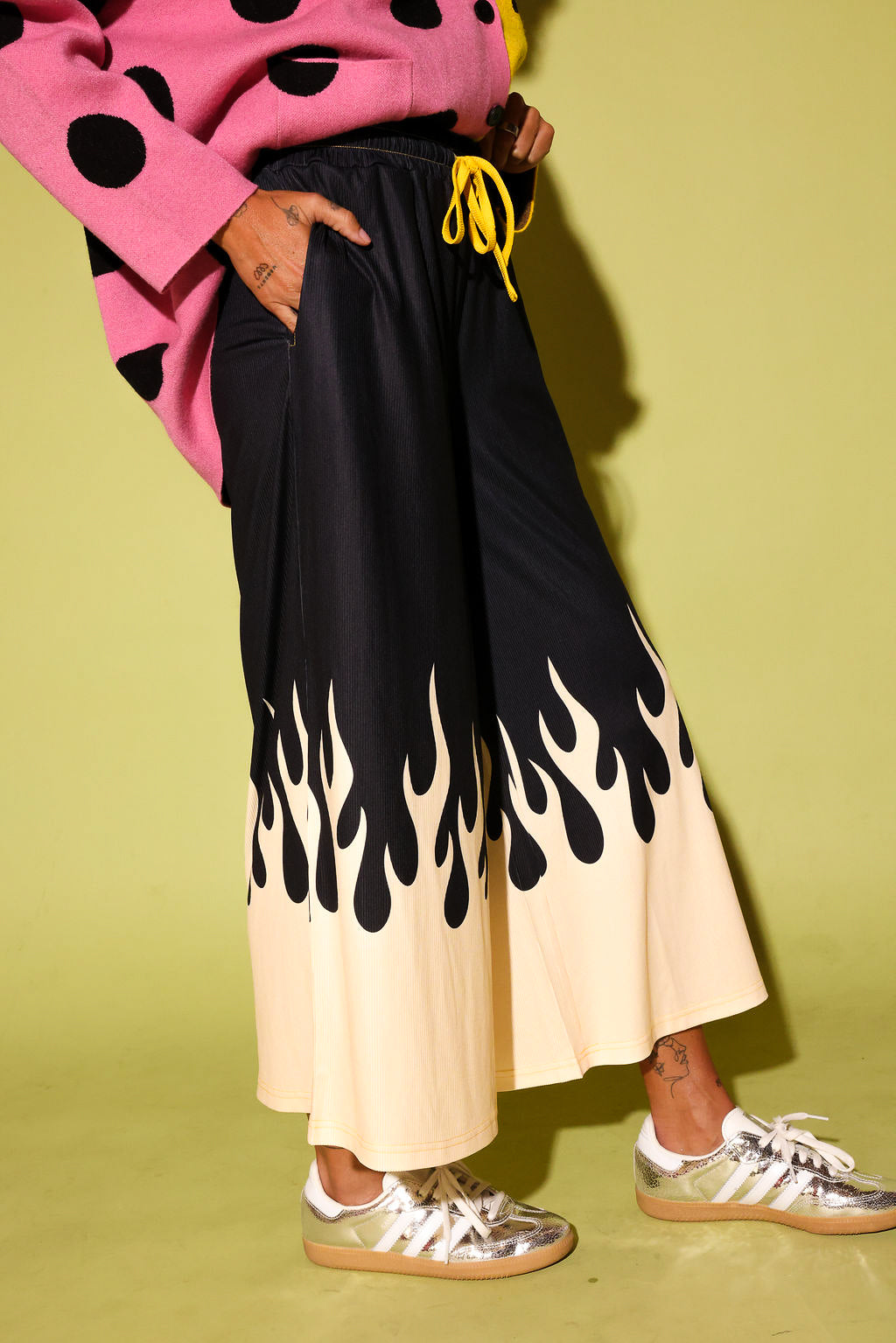 A person wearing wide-leg pleated pants with a black upper section and a cream lower section featuring a black flame print, styled with a pink polka-dot sweater and metallic sneakers against a yellow-green background.