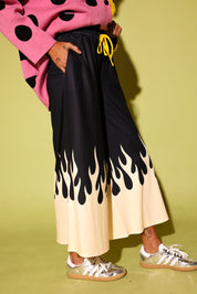 A person wearing wide-leg pleated pants with a black upper section and a cream lower section featuring a black flame print, styled with a pink polka-dot sweater and metallic sneakers against a yellow-green background.