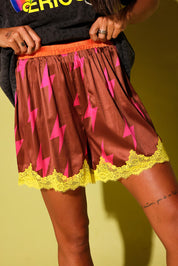 Close up of a a model wearing maroon lace shorts with pink lightening bolts and yellow lace trim holding waistband against a pale yellow background