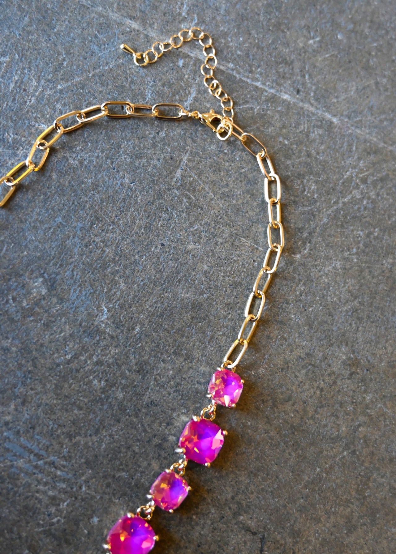 Close up of a a gold chain necklace with square neon pink gems and a clasp on a dark background