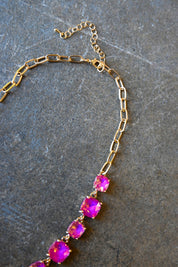 Close up of a a gold chain necklace with square neon pink gems and a clasp on a dark background