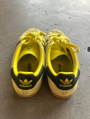 A pair of yellow and black Adidas sneakers viewed from the back, resting on a gray concrete floor. The shoes feature yellow laces, a yellow and black leather and suede upper, and a gum sole