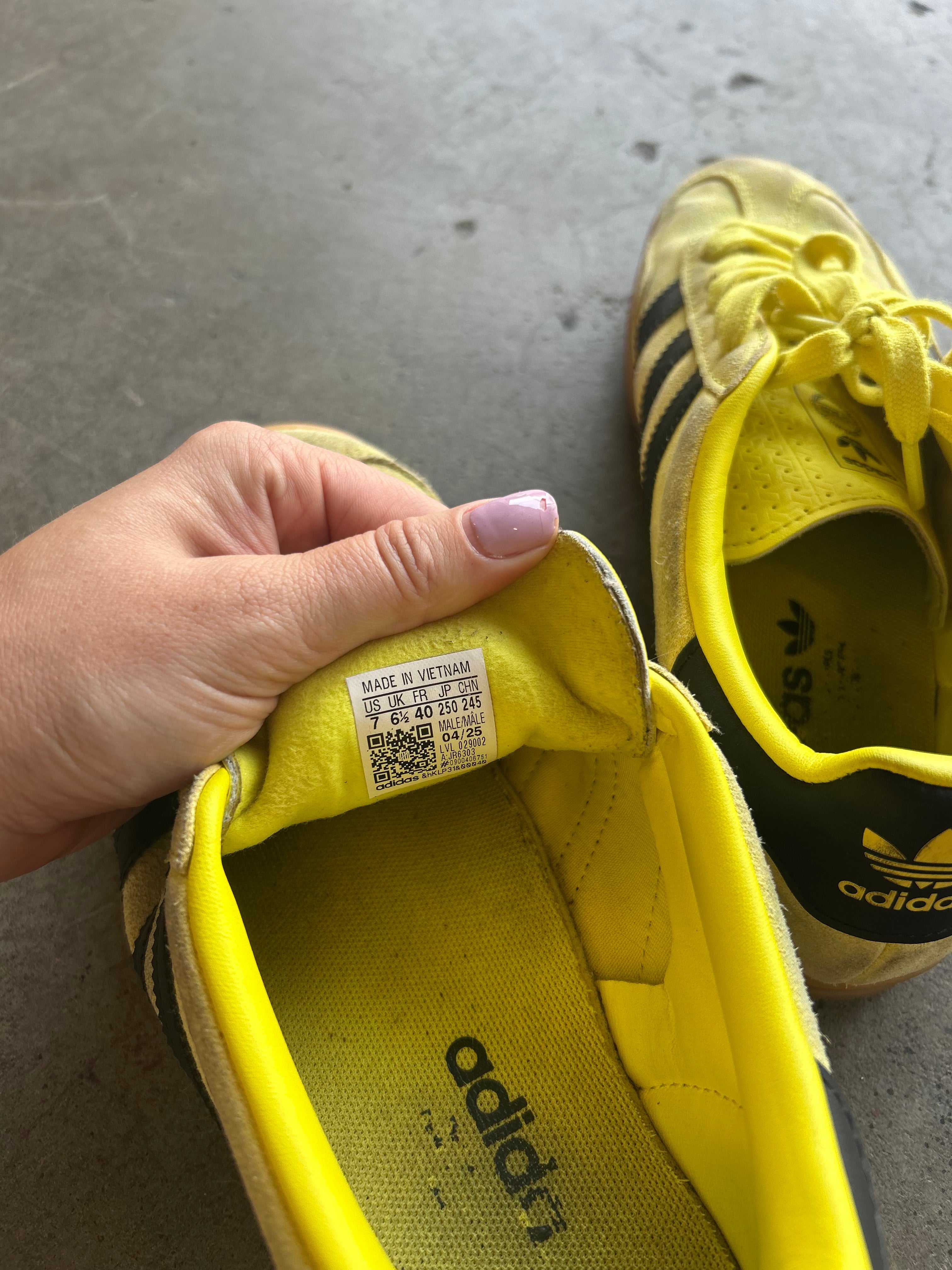 A close-up of a person's hand holding the open toe of a yellow Adidas sneaker, showing the interior label, with another yellow Adidas sneaker visible in the background on a gray concrete floor