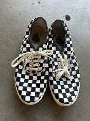 A pair of Vans checkerboard sneakers in black and white canvas with white laces, photographed from above on a gray concrete floor