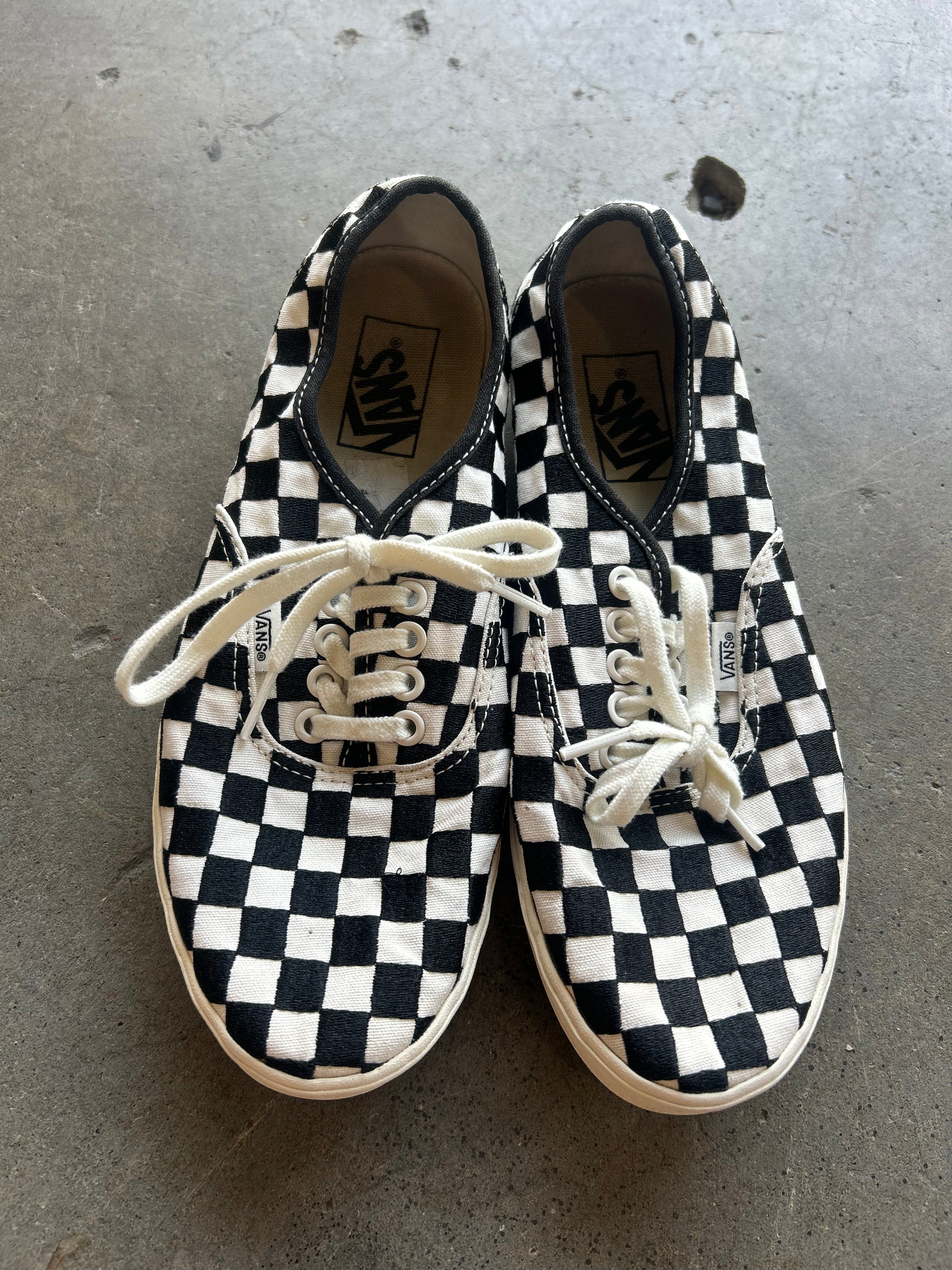 A pair of Vans checkerboard sneakers in black and white canvas with white laces, photographed from above on a gray concrete floor