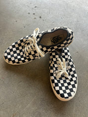 A pair of Vans checkerboard canvas sneakers with off-white laces, displayed on a gray concrete floor. The Vans logo is visible on the insole of the top shoe