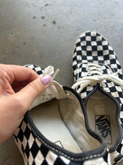 A close-up of a Vans checkerboard sneaker being held open by a hand with lavender nail polish, showing the interior lining and Vans logo, with the second shoe in the background on a gray concrete floor
