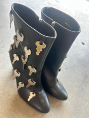 A pair of black leather knee-high boots decorated with multiple metallic keys attached along the outer shafts, photographed on a concrete floor