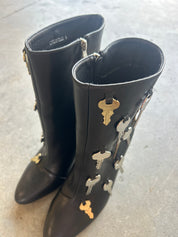 A pair of black leather mid-calf boots decorated with various metallic key charms in gold and silver, displayed on a gray concrete floor