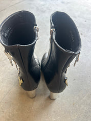 A pair of black leather ankle boots with silver metallic studs and decorative hardware, shown from the back on a concrete floor