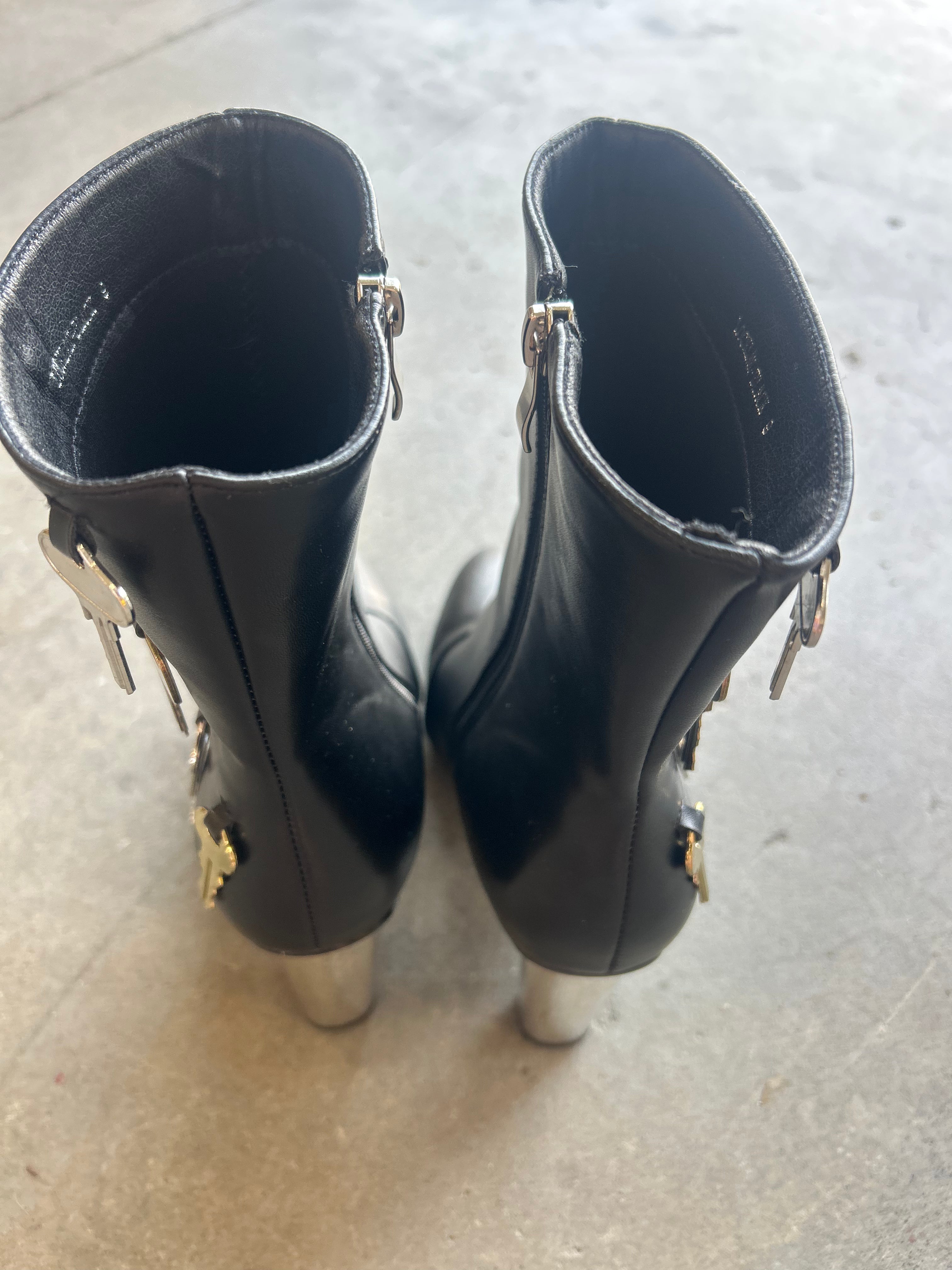A pair of black leather ankle boots with silver metallic studs and decorative hardware, shown from the back on a concrete floor