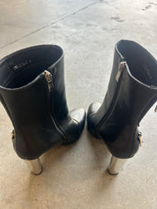 A pair of black leather ankle boots with silver metallic block heels, shown from the back on a concrete floor