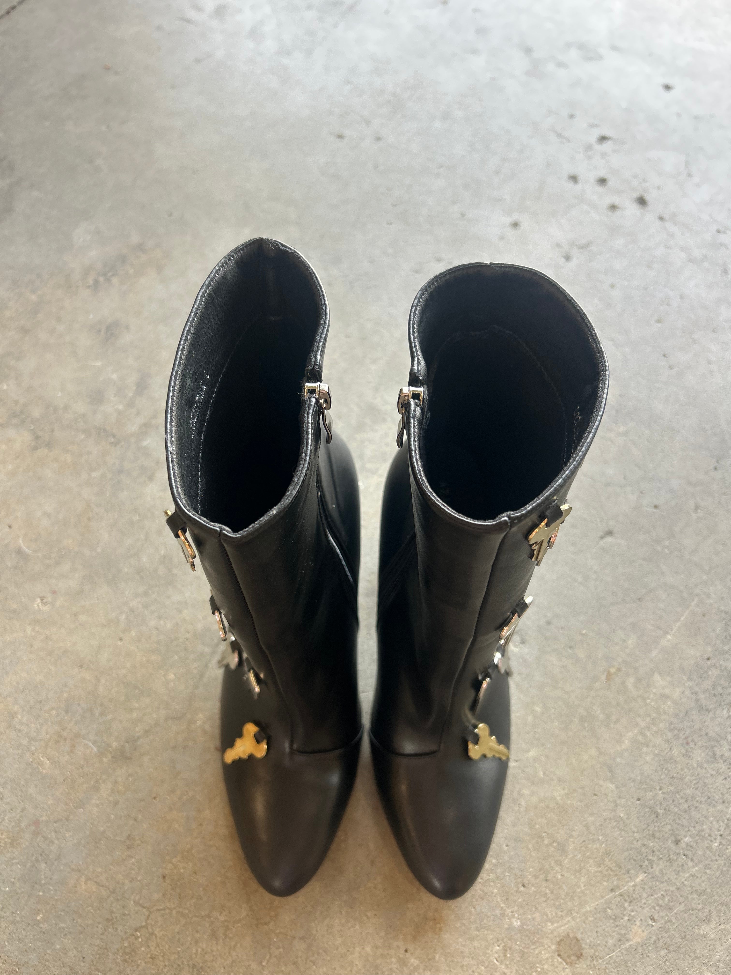 A pair of black leather ankle boots with gold-tone hardware and decorative details, photographed from above on a gray concrete floor