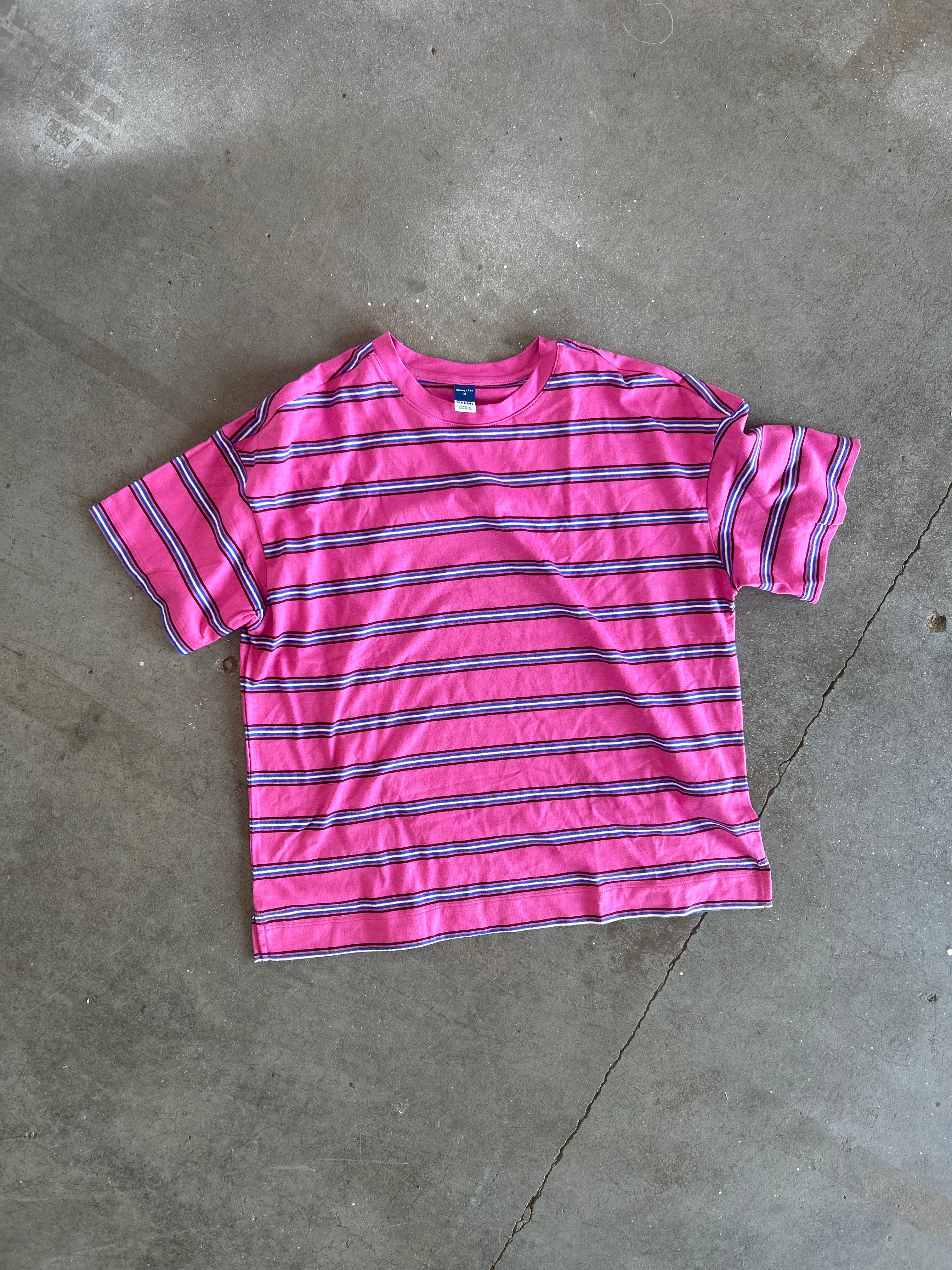 A pink short-sleeve t-shirt with horizontal navy and purple stripes, laid flat on a gray concrete floor