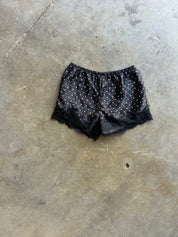 Black satin pajama shorts with white polka dots and a black lace hem, laid flat on a concrete floor