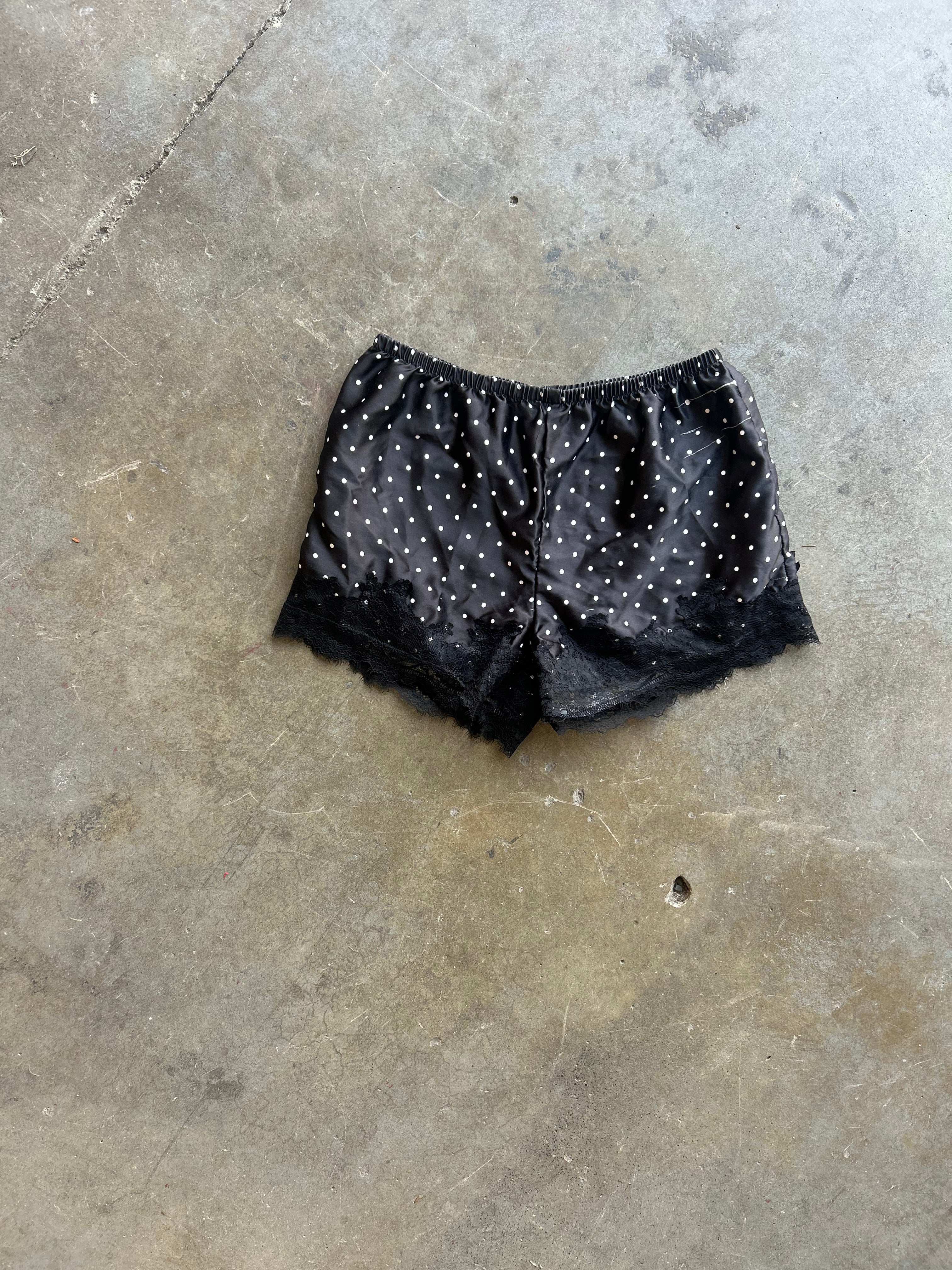 Black satin pajama shorts with white polka dots and a black lace hem, laid flat on a concrete floor