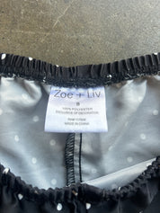 Close-up of the inside neckline of a black and white polka dot garment, showing a white size tag with the brand name 'Zoe + Liv' and black elastic trim