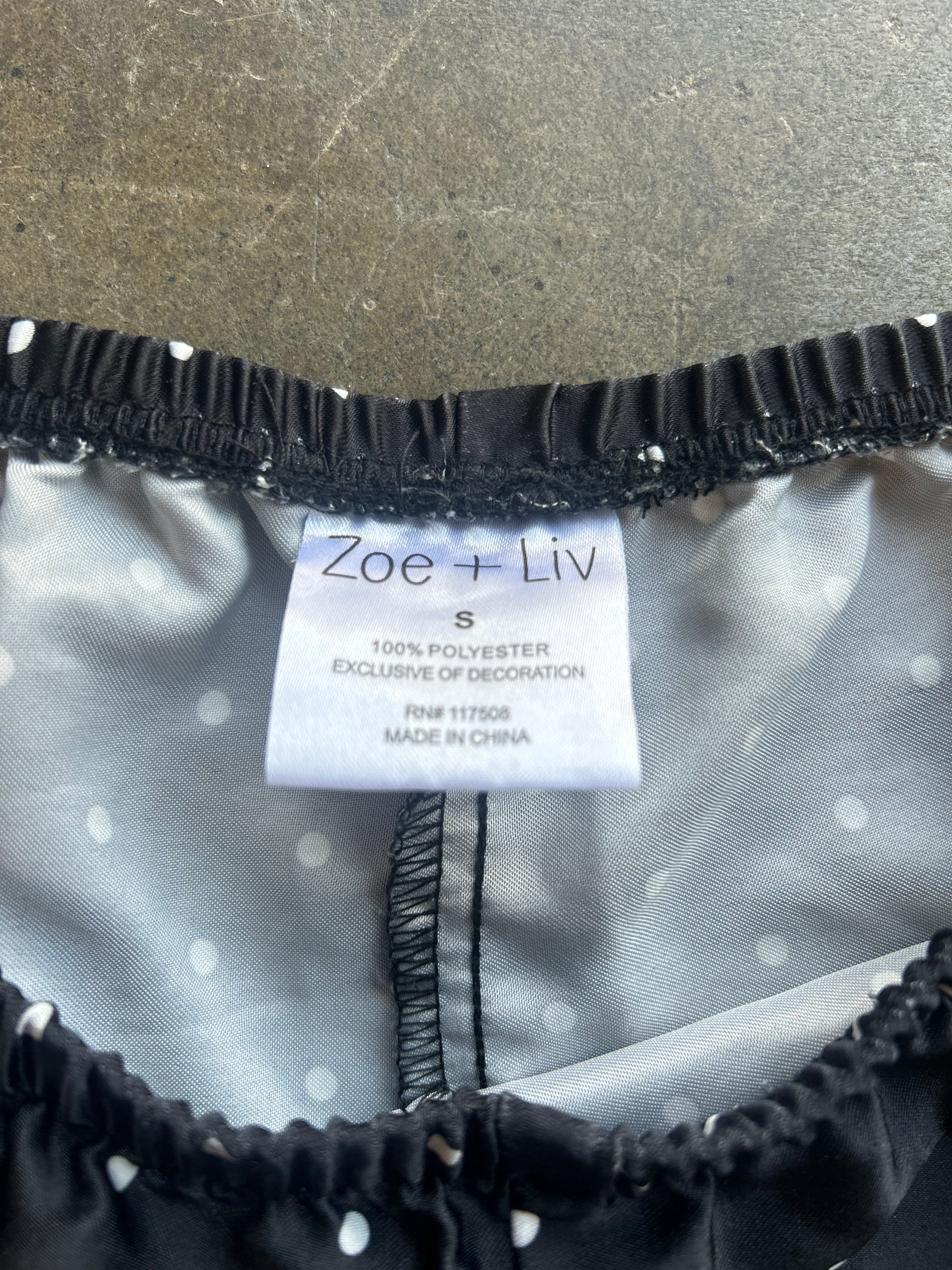 Close-up of the inside neckline of a black and white polka dot garment, showing a white size tag with the brand name 'Zoe + Liv' and black elastic trim