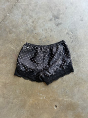 Black satin pajama shorts with white polka dots and a black lace hem, laid flat on a concrete floor