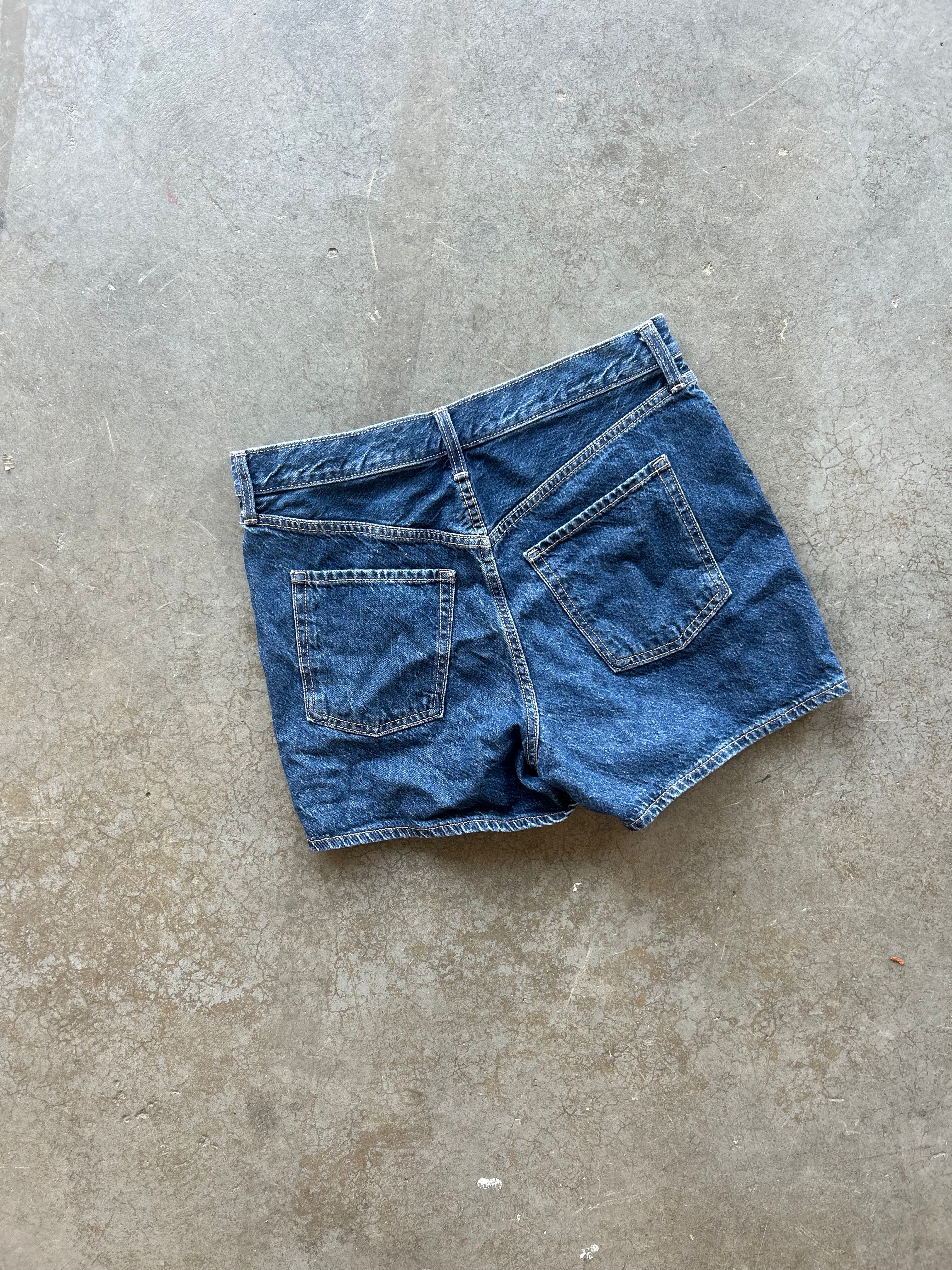 A pair of blue denim shorts shown from the back, laid flat on a textured concrete floor