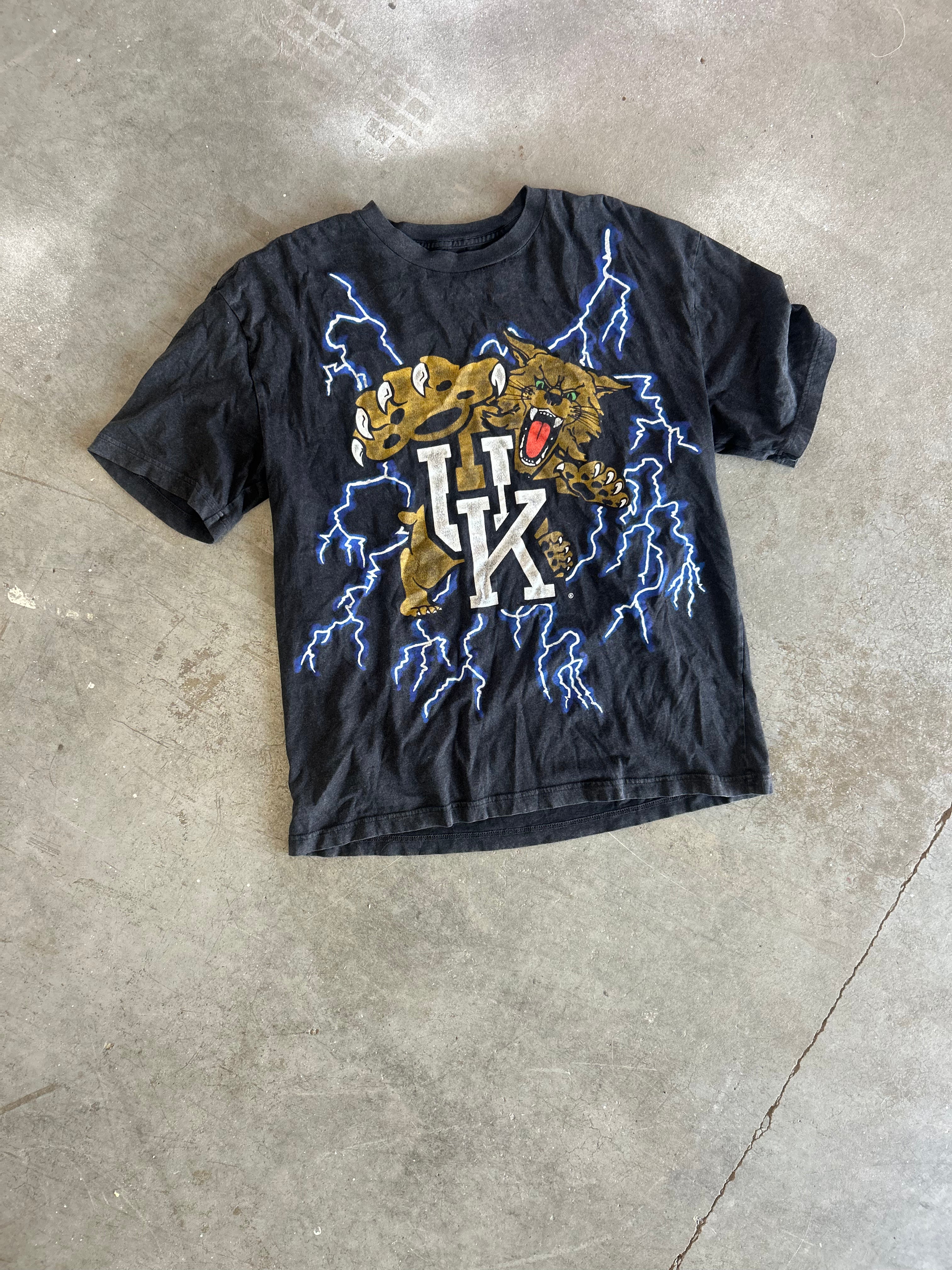 A black graphic t-shirt laid flat on a concrete floor. The shirt features a graphic of a golden-brown panther surrounded by blue lightning and a large white 'HK' monogram