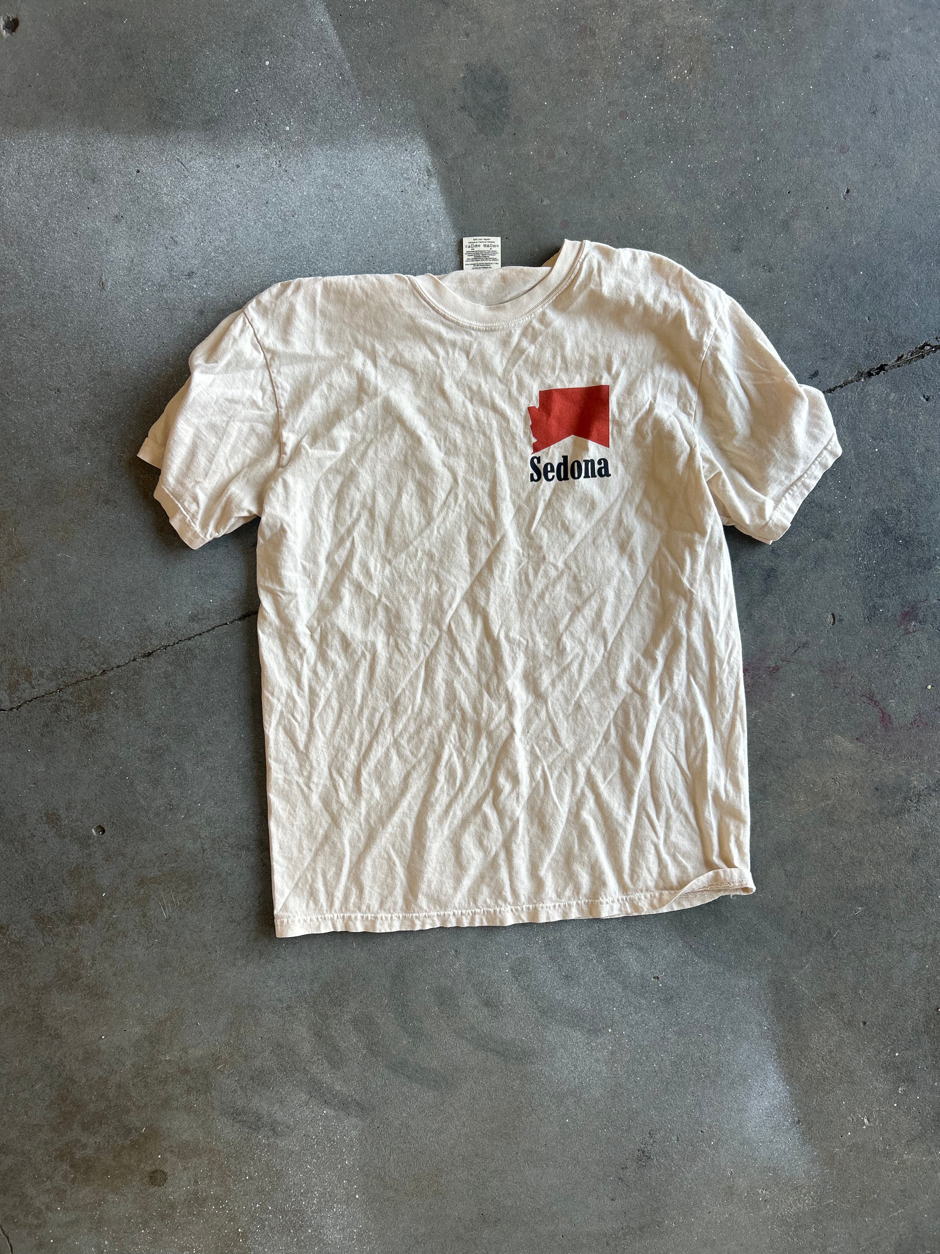 A vintage off-white short-sleeve t-shirt laid flat on a gray concrete floor. The shirt features a small red rectangular graphic on the left chest with the word 'Sedona' printed in black underneath it