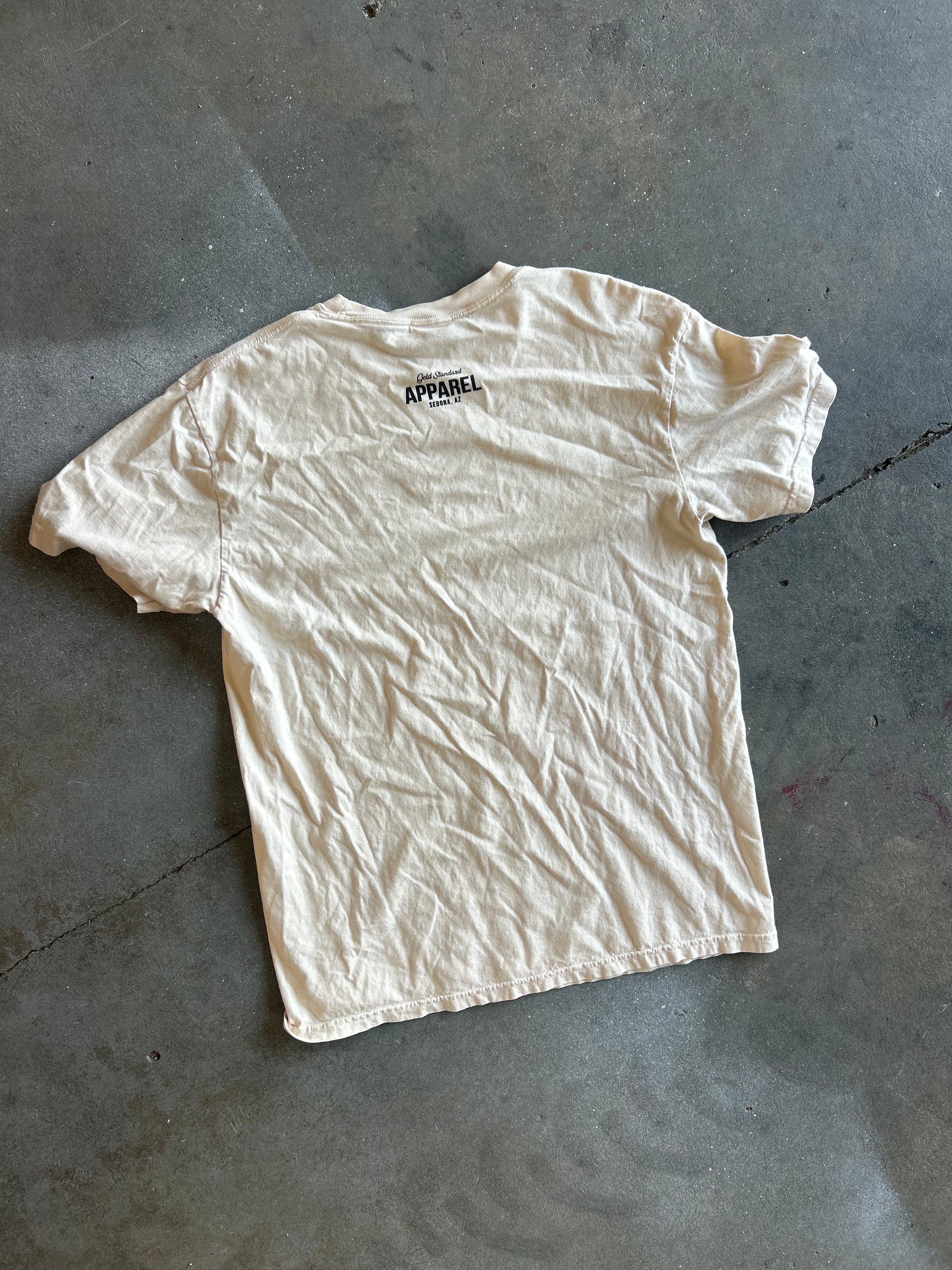 A cream-colored short-sleeve t-shirt laid flat on a gray concrete floor, featuring a small black 'APPAREL' logo printed on the upper back
