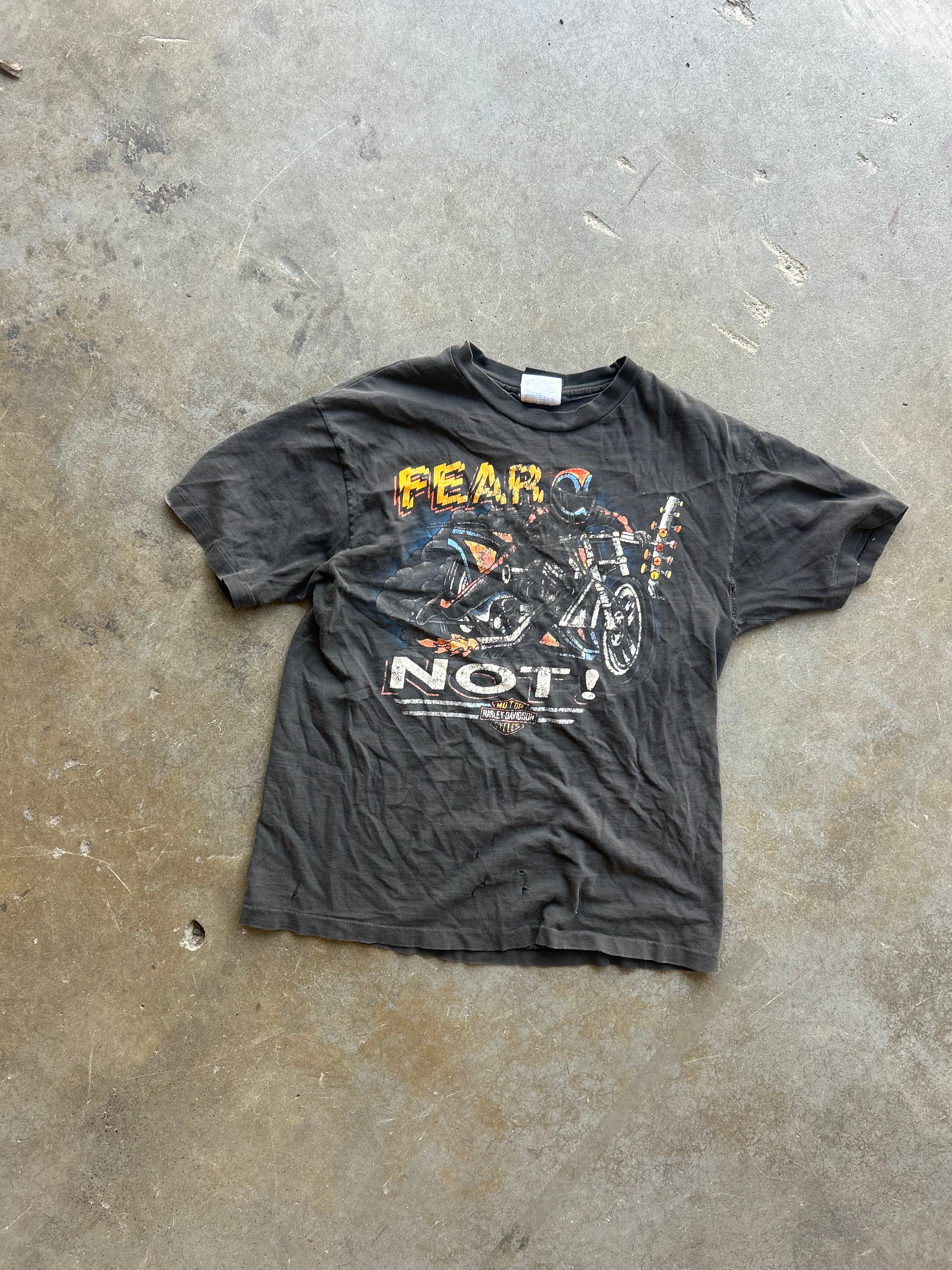 A faded black graphic t-shirt featuring a motorcycle and the words 'FEAR NOT!' in yellow and white, laid flat on a concrete floor