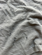 Close-up of a light gray or sage-gray fabric surface with visible wrinkles and a few small dark distressed or worn areas near the lower center