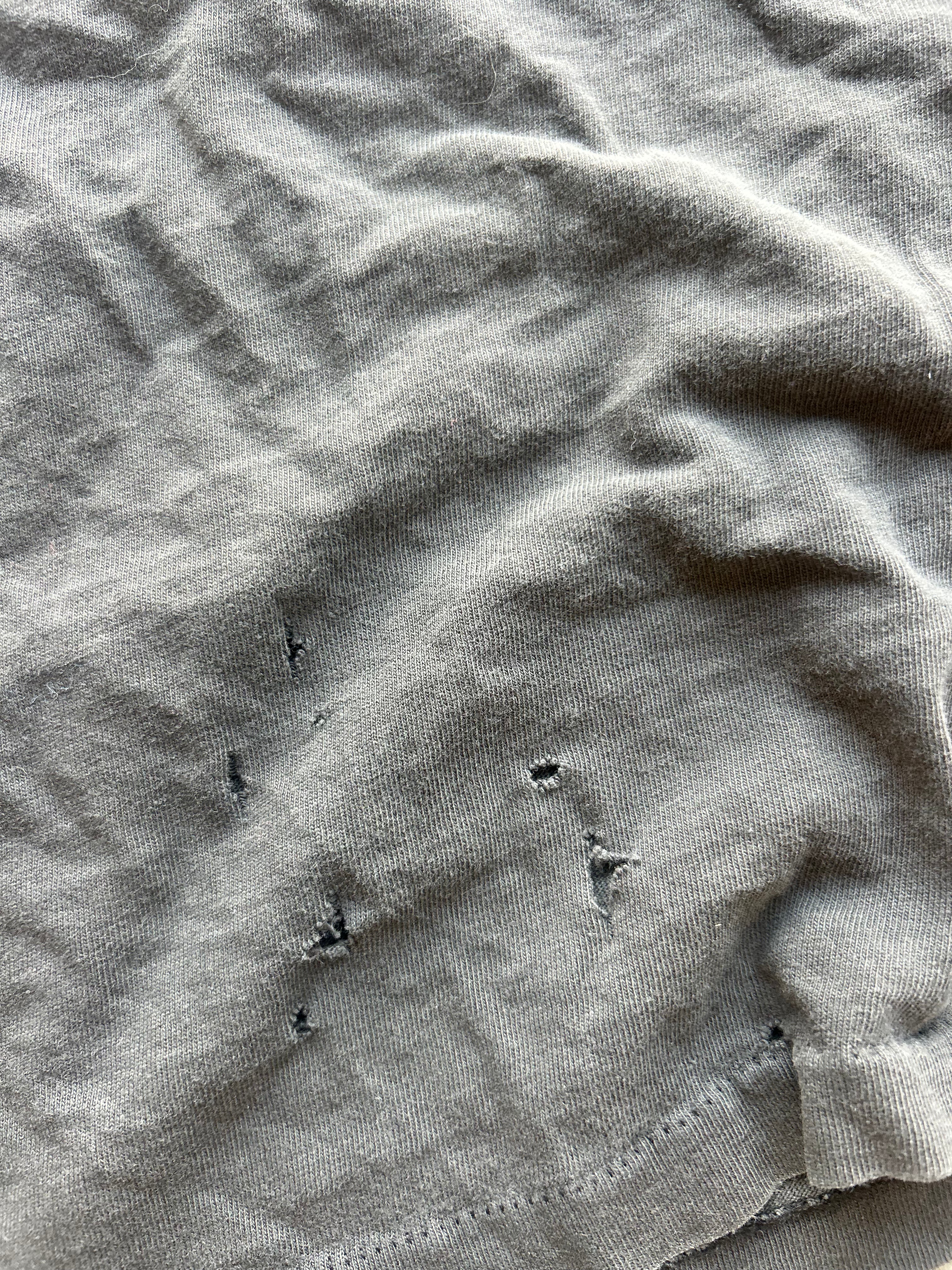 Close-up of a light gray or sage-gray fabric surface with visible wrinkles and a few small dark distressed or worn areas near the lower center