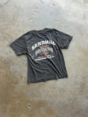A faded black graphic t-shirt featuring a vintage-style print of Harley-Davidson and Sandhill, laid flat on a concrete floor