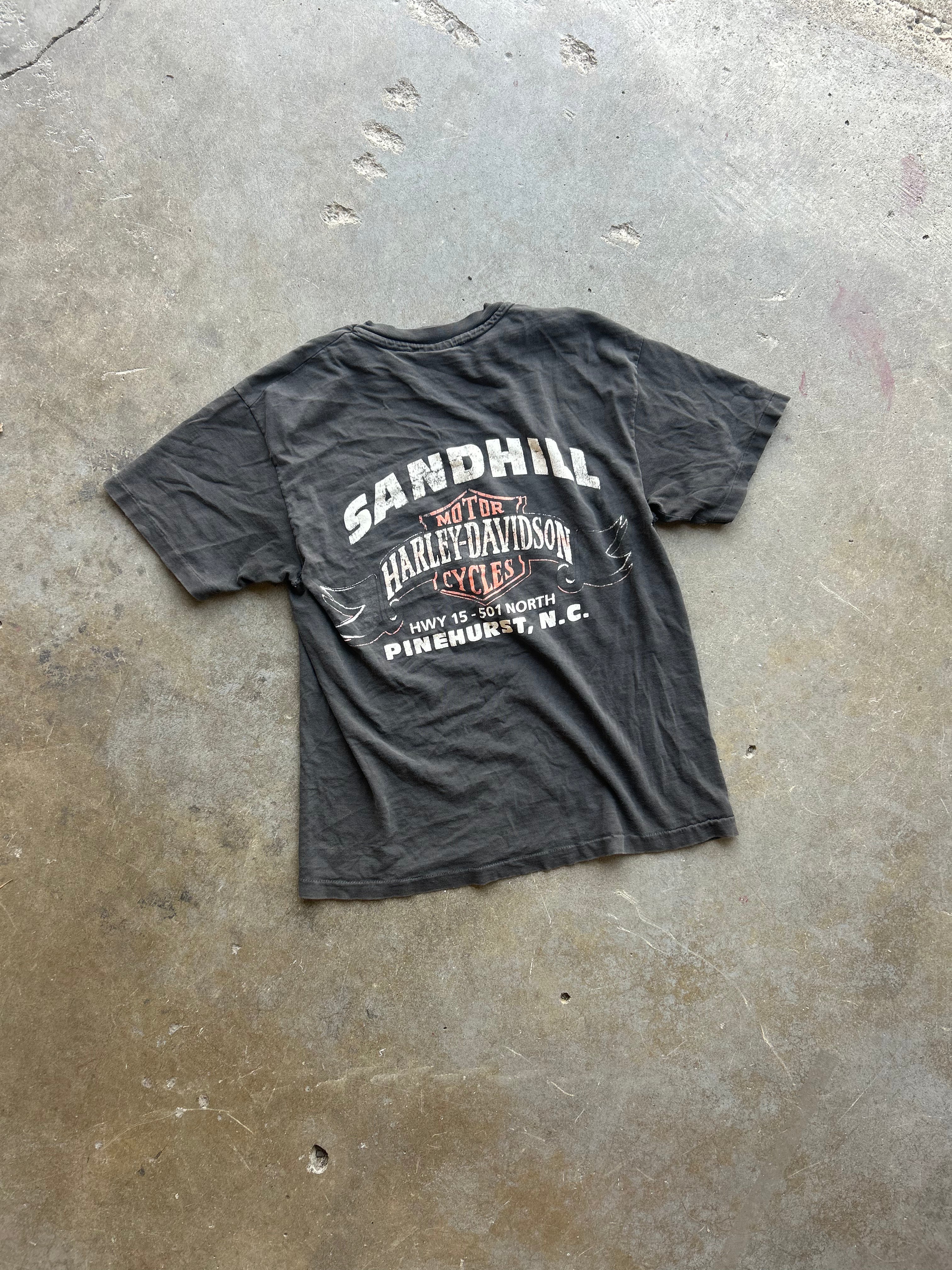 A faded black graphic t-shirt featuring a vintage-style print of Harley-Davidson and Sandhill, laid flat on a concrete floor