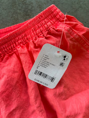 A close-up of bright coral pink shorts with an elastic waistband and a white retail price tag attached. The tag shows text including 'C