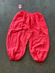 Bright red crinkled cotton harem-style pants with elastic waistband and drawstring ankle cuffs, laid flat on a dark gray textured surface