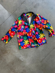 A long-sleeve button-up jacket with a vibrant, multicolor floral print, laid flat on a gray concrete floor. The jacket features a mix of red, yellow, blue, green, and pink flowers on a bright blue background, with a black collar decorated with white polka dots