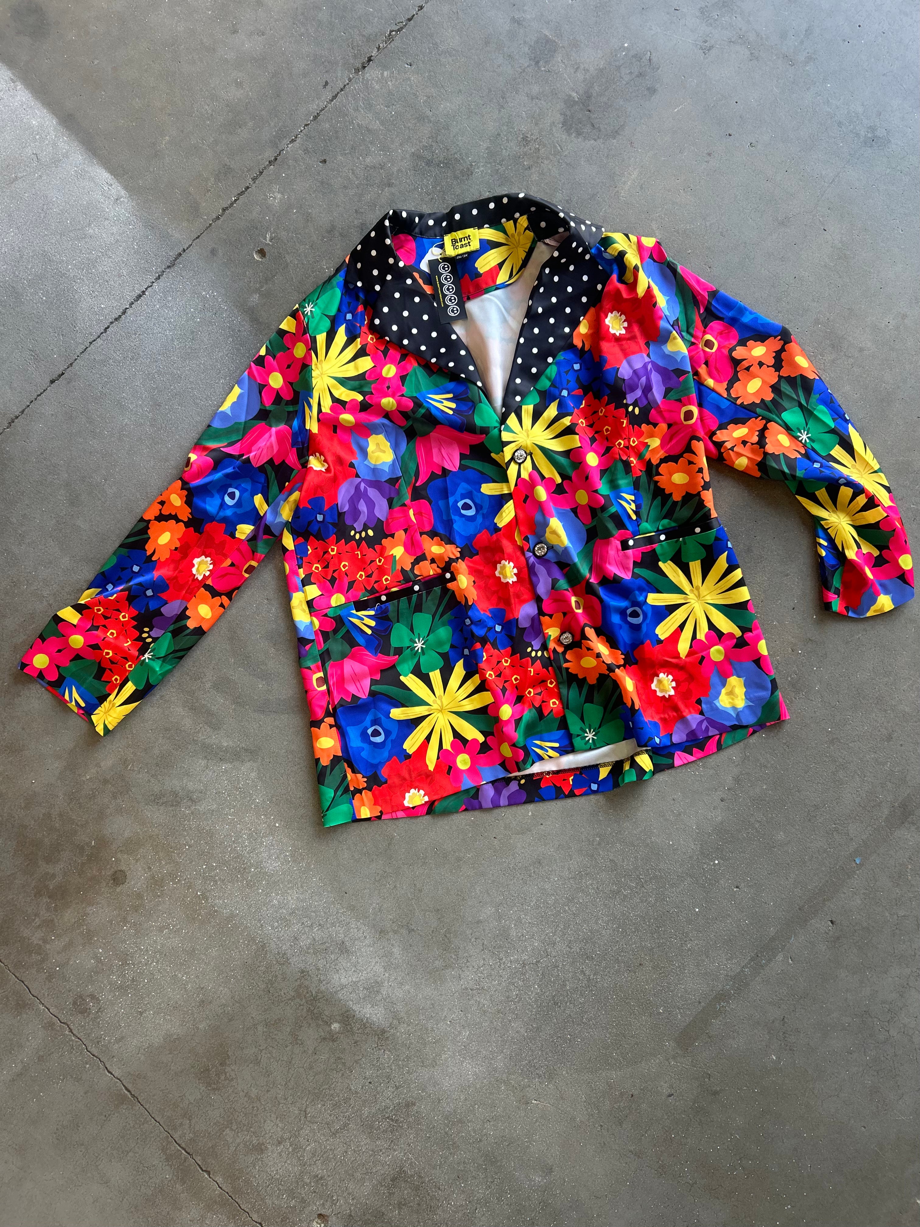 A long-sleeve button-up jacket with a vibrant, multicolor floral print, laid flat on a gray concrete floor. The jacket features a mix of red, yellow, blue, green, and pink flowers on a bright blue background, with a black collar decorated with white polka dots