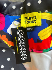 Close-up of a colorful garment with a black and white polka dot pattern on one side and a bright abstract print in blue, yellow, red, orange, and green on the other. A yellow Burnt Toast brand tag and a black hangtag are attached