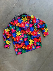 A long-sleeve collared shirt with a vibrant, multicolor floral print, laid flat on a gray concrete floor