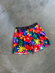 A pair of colorful floral print shorts with a black waistband featuring white polka dots, laid flat on a gray concrete floor