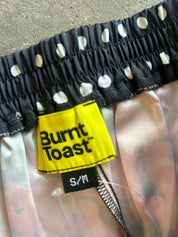 Close-up of a garment waistband with a black elastic band featuring white polka dots, a bright yellow Burnt Toast brand label, and light pink patterned fabric below