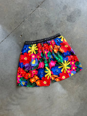 A pair of colorful floral shorts with a black waistband, laid flat on a gray concrete floor