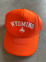 Bright orange trucker hat with white embroidered 'WYOMING' text and a white bucking horse logo on the front, photographed on a gray concrete surface