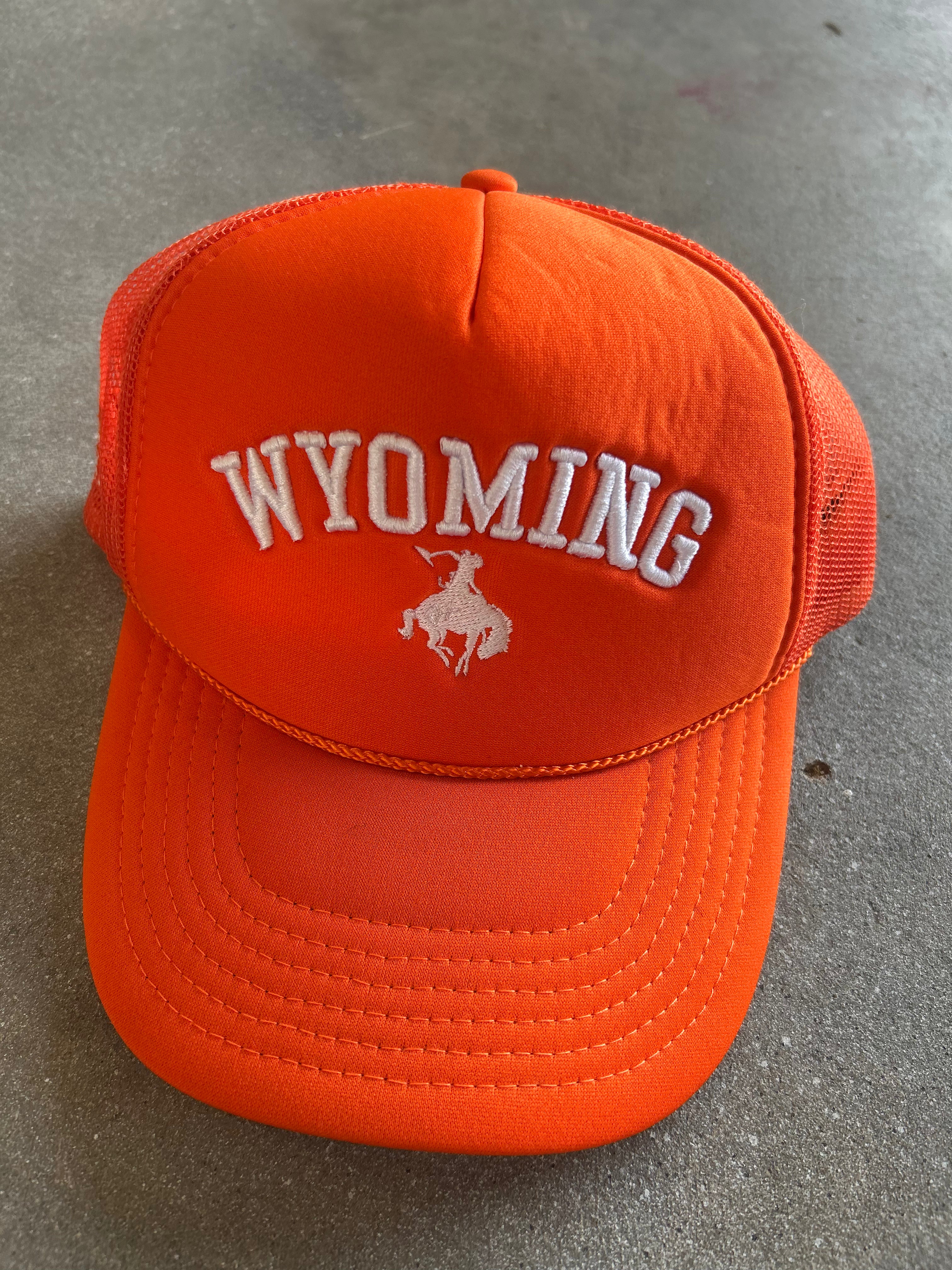 Bright orange trucker hat with white embroidered 'WYOMING' text and a white bucking horse logo on the front, photographed on a gray concrete surface