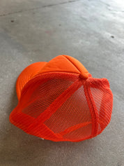 An orange trucker hat with a solid orange front panel and matching orange mesh back, photographed on a gray concrete floor