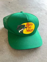 A green Bass Pro Shops trucker hat with a yellow oval logo and fish graphic on the front, resting on a gray concrete surface