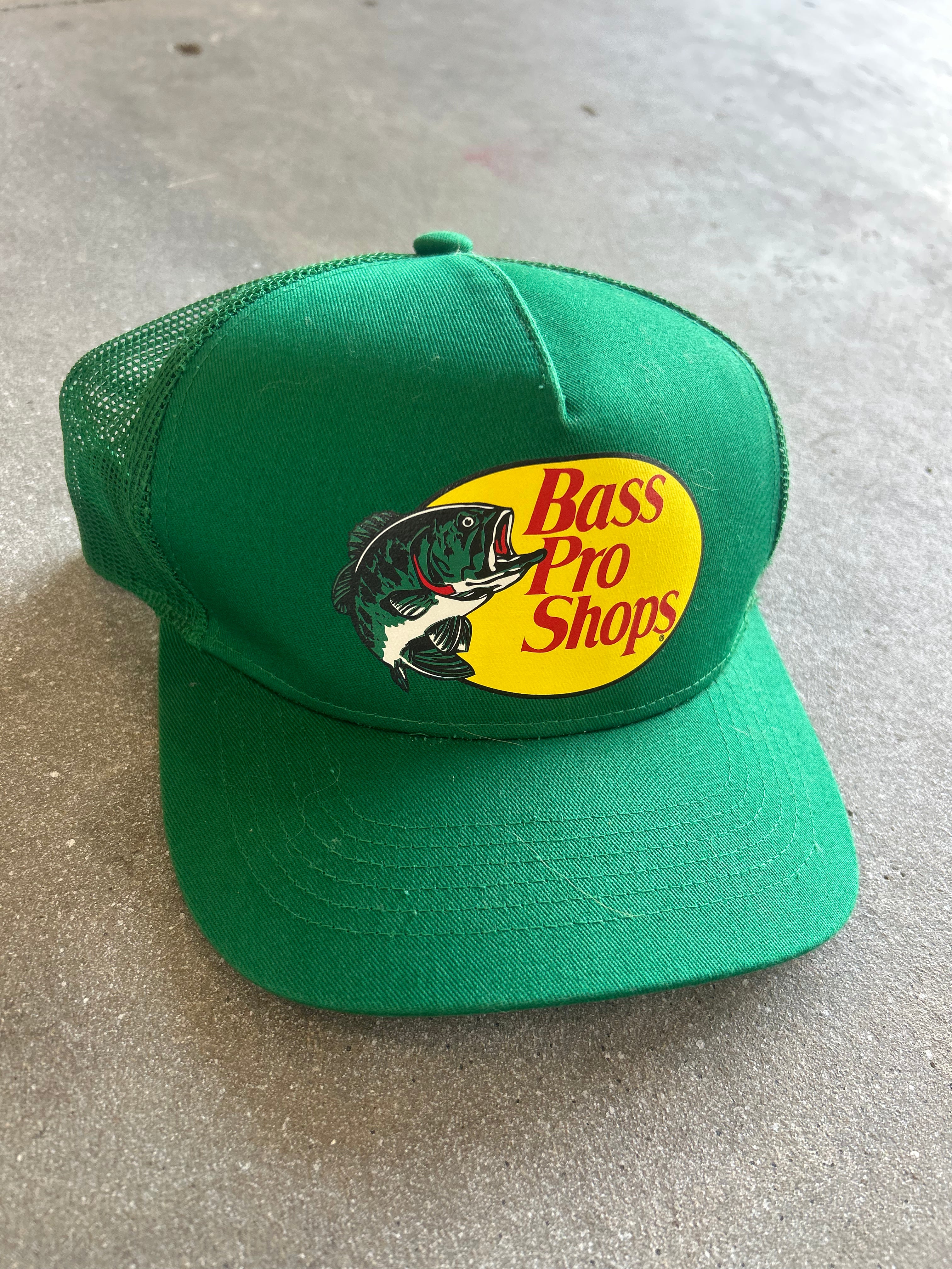A green Bass Pro Shops trucker hat with a yellow oval logo and fish graphic on the front, resting on a gray concrete surface