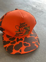 An orange and brown camouflage trucker hat with a black embroidered 'Leroy Brown' logo on the front, resting on a gray concrete floor