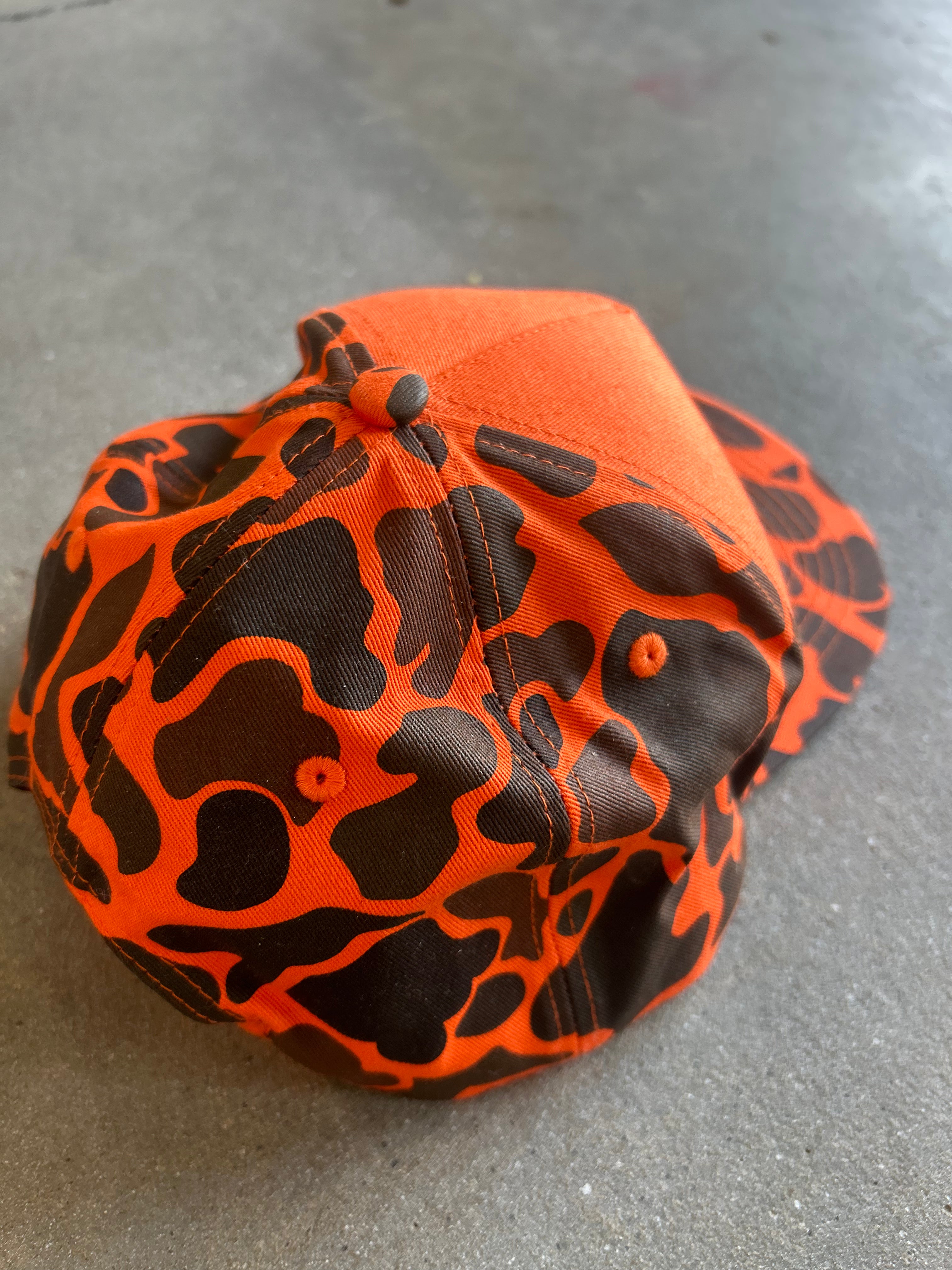A bright orange and black camouflage-patterned baseball cap resting on a gray concrete floor
